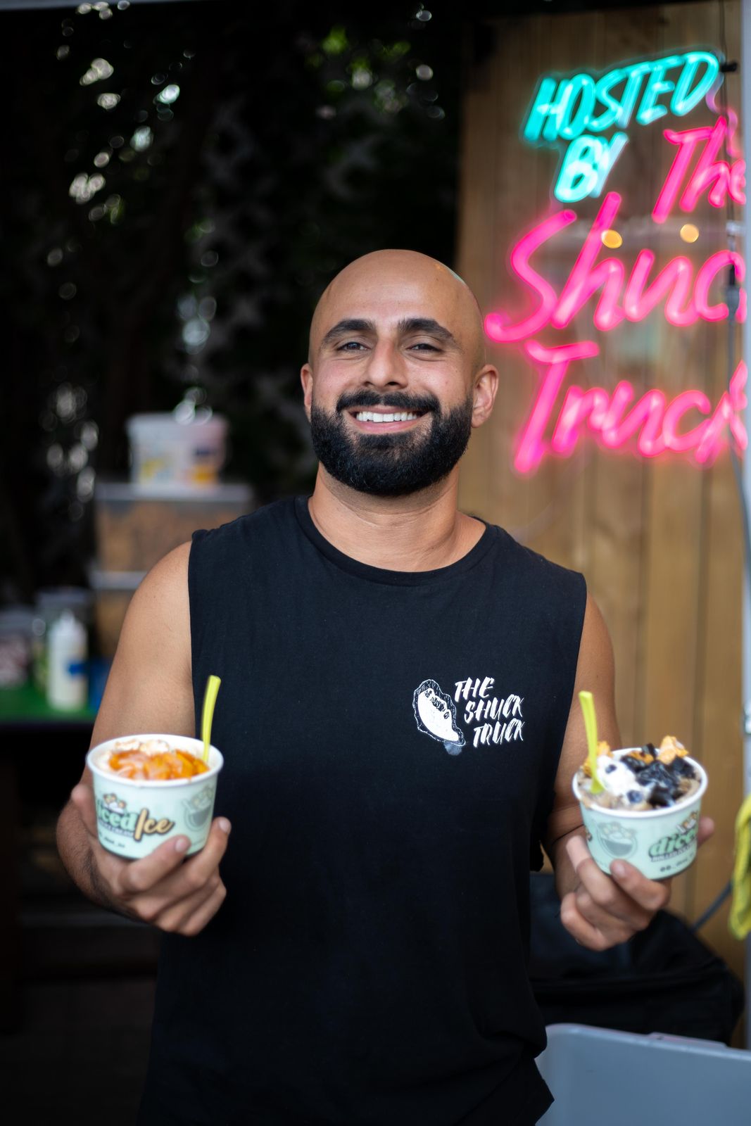 Holding two drinks under the pink Hosted by The Shuck Truck neon sign at Art Crawl