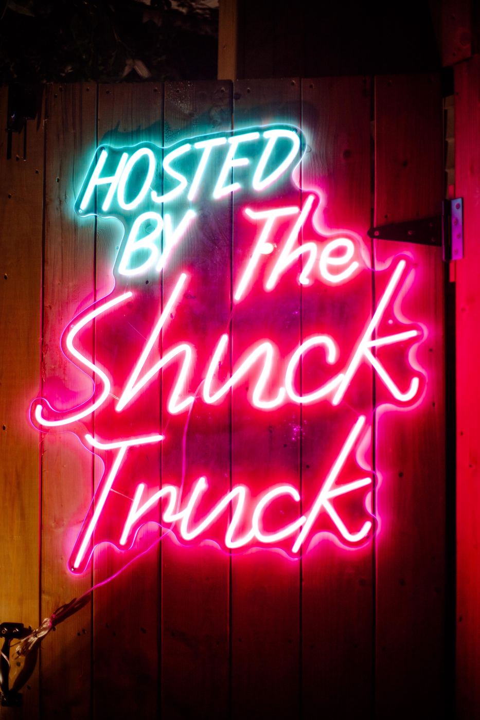 The pink neon Hosted by The Shuck Truck sign on a wooden wall
