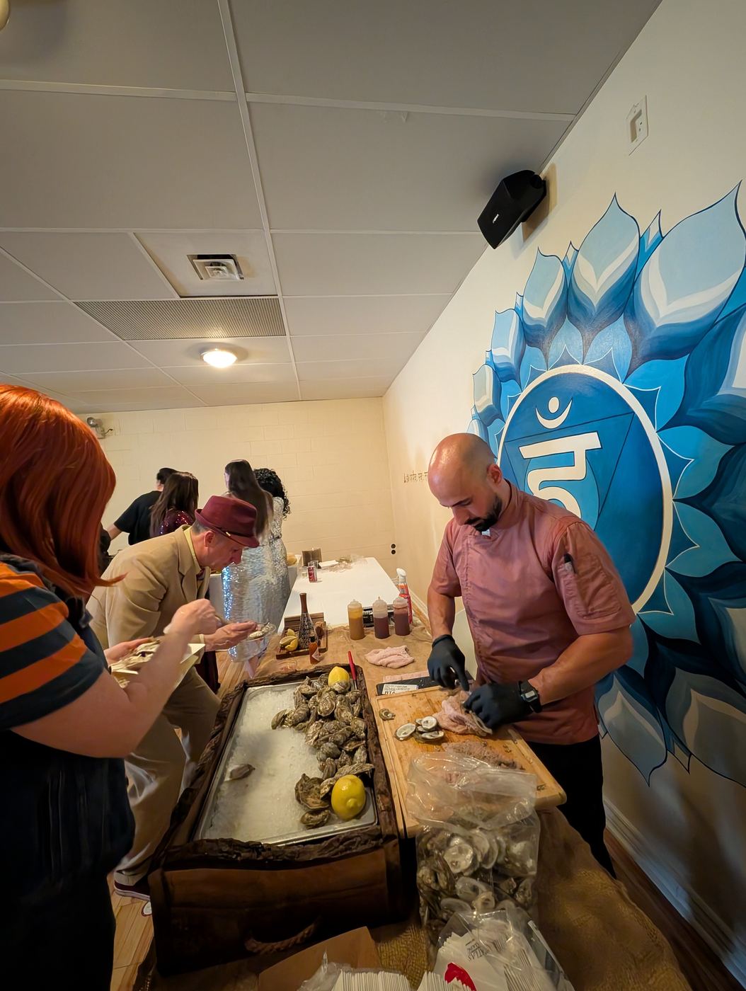 Ramsen shucking oysters at the live-edge box in front of a blue mural while guests eat